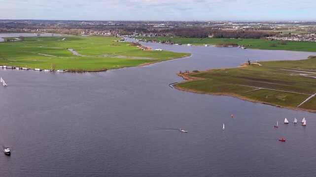 High angle aerial drone video clip of sailboats cruising on wide lake basin, green polder fields, traditional windmill, houseboats, and distant town horizon in Netherlands, West Europe