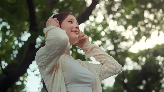 Happy young Asian woman putting on wireless headphones and gently moving to music outdoors, joyful lifestyle and relaxation concept