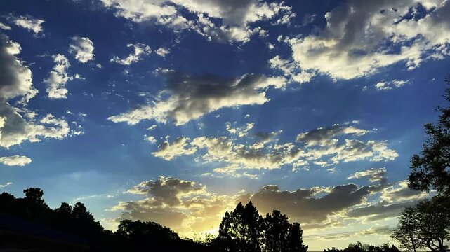 A stunning view of the evening sky at sunset. Golden light and sunbeams (crepuscular rays) pierce through masses of white clouds drifting slowly across a clear blue sky.
