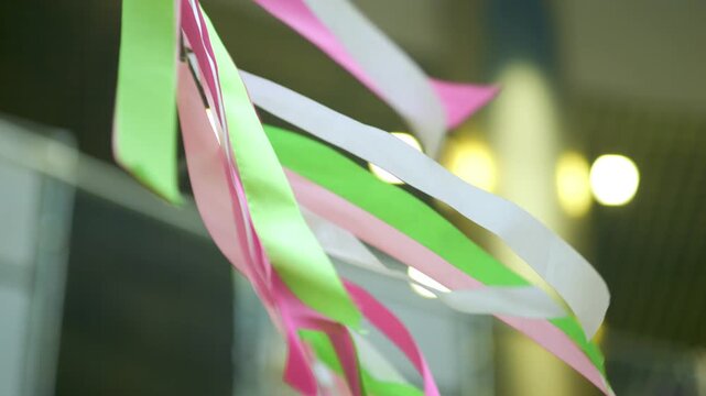 Colorful pink green white ribbons sway gently in soft breeze. Blurred warm lights suggest indoor celebration space. Festive energy fits party promotions or event decor