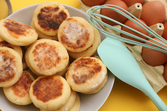 Homemade delicious syrniki, cheese pancakes in plate, cooking ingredients, kitchen utensils on yellow background