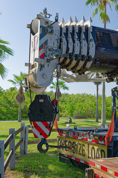 Heavy-duty crane hook and telescopic boom secured on a flatbed truck trailer displaying an oversize load sign, set outdoors near palm trees and green landscaping on a sunny day.
