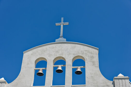 Three church bells suspended in arched openings of a white stucco bell tower against a vivid blue sky, suggesting tradition, calm and sunlit Mediterranean architecture with clean minimalist lines.
