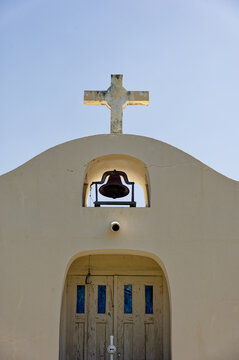 Sunlit white chapel facade featuring a weathered cross and bell set above rustic wooden doors with blue glass insets, evoking serenity, tradition, and Mediterranean or Southwestern architectural chara
