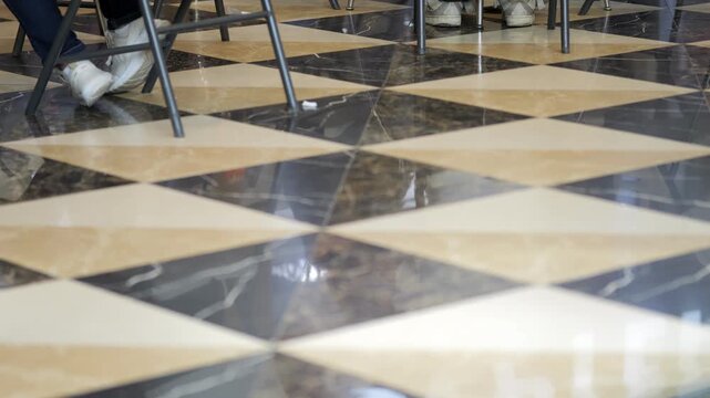 The black and beige checkered marble floor shines underfoot. People's legs move through the patterned tiles. Light jeans and casual footwear create dynamic foot traffic