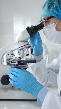 Female scientist wearing a mask and blue safety gear, using a microscope for research while a male colleague works in the background. Medicine, healthcare and science concept