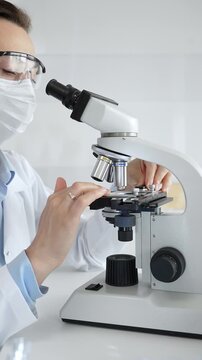 Female scientist examining specimen under microscope, conducting research in sterile laboratory with colleague working near test tube rack. Medicine, healthcare and science concept