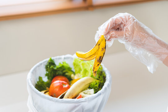 Hand in plastic glove throwing a banana peel into a trash bin filled with food waste and vegetable scraps, illustrating organic waste separation and eco friendly composting concept.