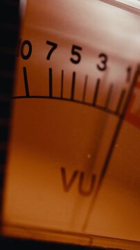 Illuminated analog VU meter with mechanical needle moving across decibel level scale on vintage sound equipment. Vertical macro shot