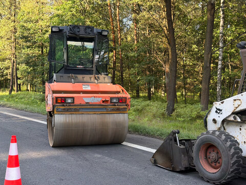 Road roller working on a paved road near a forest with a traffic cone and construction vehicle parts