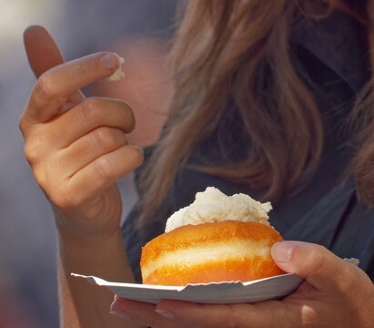 A woman picks a bite of whipped cream from a golden fried pastry bun on a paper plate at Naplavka farmers market in Prague, her face out of frame in warm afternoon sunlight.

