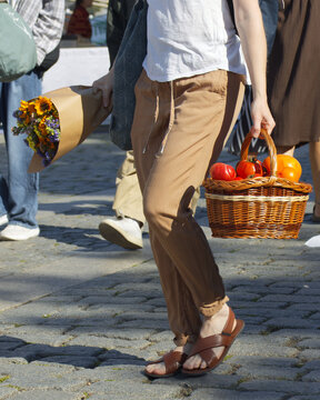 A shopper strolls across cobblestones at Naplavka farmers market in Prague, carrying a paper-wrapped sunflower bouquet and a woven basket of heirloom tomatoes in warm sunlight.