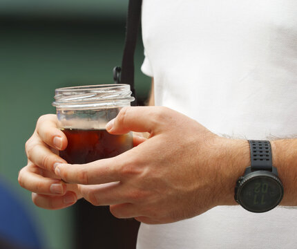A close-up of a hand holding a glass jar filled with dark coffee at Naplavka farmers market in Prague, with a sports watch and camera strap visible.