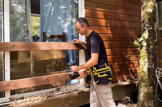 man on an extension ladder using a hammer to nail in new wood siding boards, homeowner maintenance and upkeep, making outdoor repairs on house 