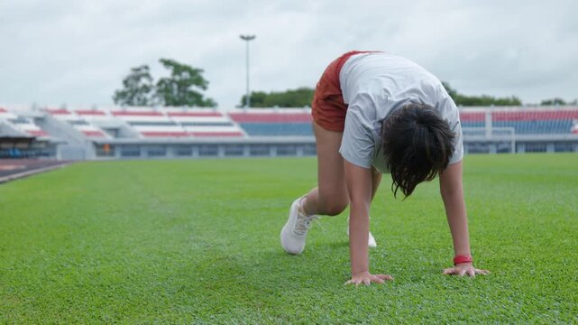 Authentic shot of an active Asian woman practicing a stretch on a beautiful green soccer field. The empty stadium with colorful seats provides excellent copy space for sports backgrounds.