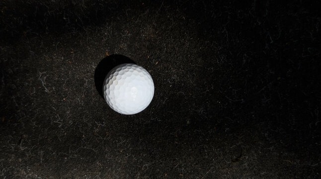 Golf ball resting on ground, in dark textured surface, with golf ball, from a top-down perspective perspective, with white, black accents, and stillness mood, with nearby objects.