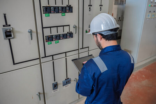 Focused professional male industrial electrical engineer safely uses a digital tablet to inspect a large power control panel during maintenance inside a modern factory manufacturing plant.