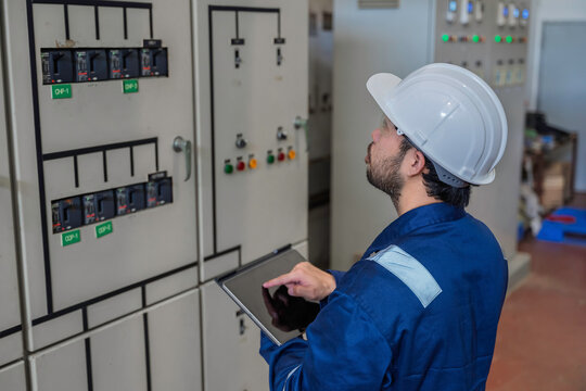 Focused professional male industrial electrical engineer safely uses a digital tablet to inspect a large power control panel during maintenance inside a modern factory manufacturing plant.