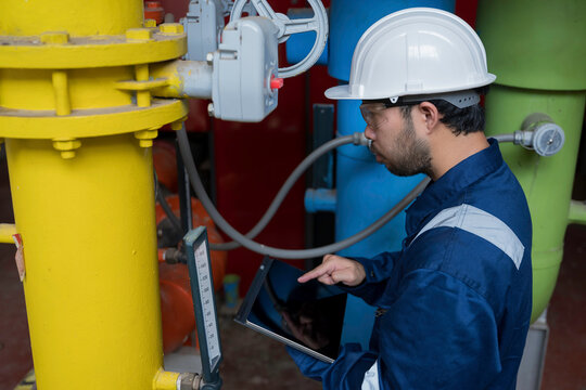 Focused professional male industrial engineer uses a laptop computer to inspect heavy machinery and colorful utility pipes during maintenance inside a modern manufacturing plant facility.