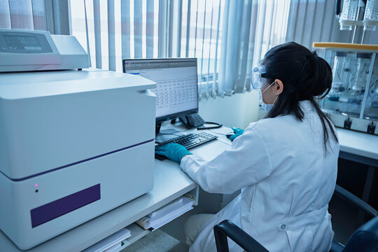 Female researcher analyzes experimental data on a computer beside a microplate reader in a laboratory. Concept of scientific research, biotechnology, diagnostics, and laboratory analysis.
