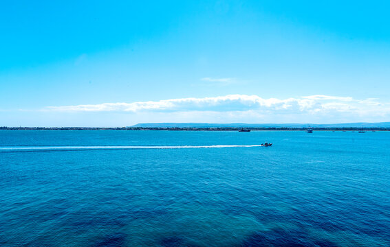 A sweeping panoramic view of the blue Sicilian sea, crisscrossed by the wake of a small boat beneath a clear sky with a streak of luminous clouds on the horizon
