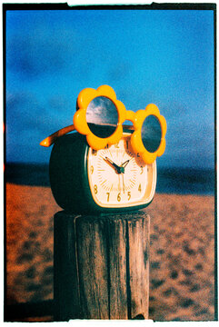 flower-shaped sunglasses rest on an alarm clock at the beach, 35mm
