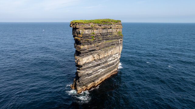 Aerial view of a towering sea stack in Ireland with layered rock cliffs rising from blue water of the Atlantic Ocean beneath a clear sky.