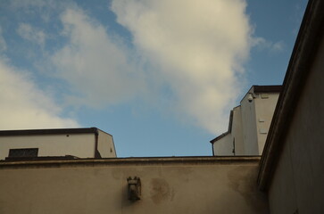 Low angle view of historic stone and plaster building facades featuring a small sculpted stone corbel and a modern security camera against a blue sky with clouds in Sicily, Italy. © beya