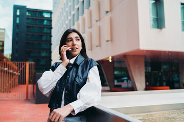 Fototapeta premium Spanish female student phone calling looking away standing. Hispanic woman traveler talking cellular device conversation mobile operator roaming network near contemporary glass building facade.