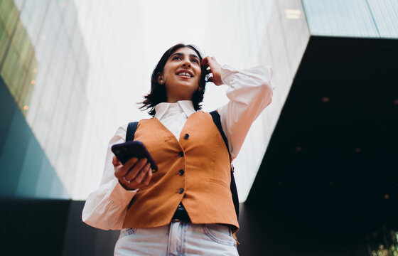 Happy hispanic woman student touch hair browsing mobile message near modern building. Smiling latin female tourist checking map navigation route online application program look away.