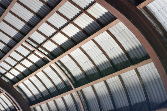 An architectural low angle view of a curved warehouse roof featuring corrugated metal panels supported by a framework of dark steel beams.