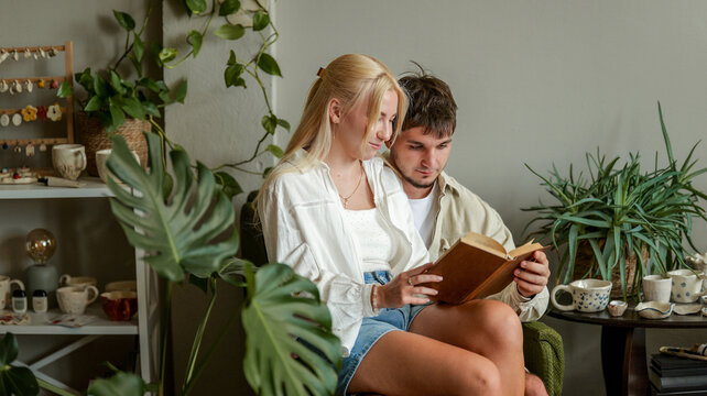 Young couple reading a book together in a cozy room filled with plants and pottery