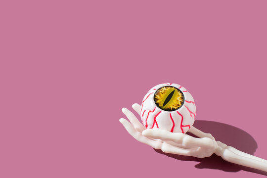 Human skeleton hand holing eyeball with bloody vein on a bright purple background. Halloween concept.