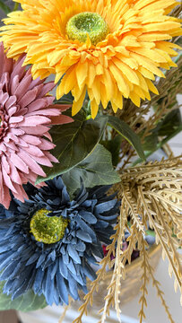 Colorful artificial flower arrangement with yellow, pink and blue blossoms, green leaves and decorative foliage, close-up floral composition for interior decor.