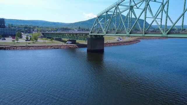 Aerial underpass showing the underside of the J.C. Van Horne Bridge as the drone glides near a concrete pier, with Sugarloaf Mountain and passing vehicles completing the Campbellton landscape.