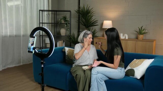 Young Asian influencer filming a beauty tutorial applying makeup on her caucasian grandmother