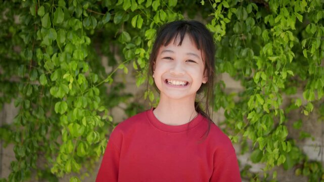 Portrait of adorable Asian kid looking directly at the camera with a gentle smiling face in a red shirt standing outdoors with beautiful green bokeh background. Childhood happiness lifestyle concept.