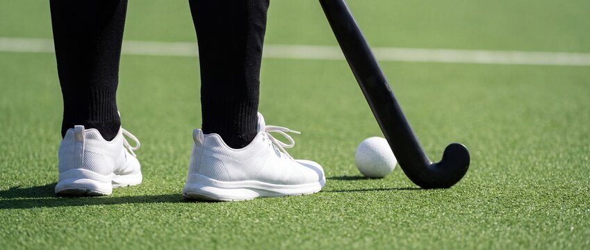 Field hockey player standing ready with stick and ball on synthetic green turf sports pitch with positioned next black stick white ball green field wearing white sneakers