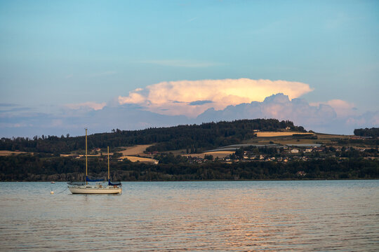 Petit voilier sur le lac de Neuch&acirc;tel avec des collines en arri&egrave;re-plan et de grands nuages &eacute;clair&eacute;s par la lumi&egrave;re du soir.