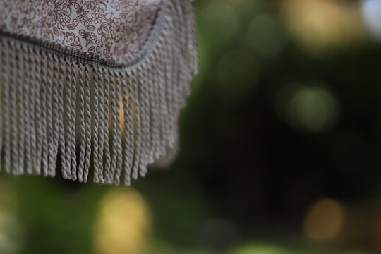 The image shows a close-up of fringe on a textile hanging in a garden. Sunlight filters through trees, creating a soft background. The scene captures elements of nature and fabric.