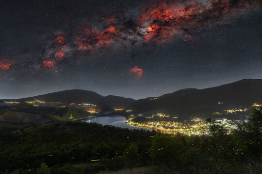 View of Lago di Fiastra at night with the Milky Way and vibrant nebulae in the starry sky above the illuminated town and surrounding hills in Fiastra, Marche, Italy.