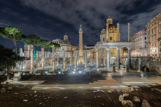 View of Trajan's Forum at night, featuring the illuminated Trajan's Column, ancient marble columns, and historic ruins set against a cloudy sky, Roma, Lazio, Italy.