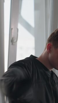 Young man with short brown hair puts on black zip up jacket. Man adjusts garment over his dark shirt maintaining focus. Scene is bright and modern featuring white walls
