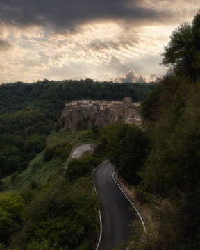 View of the historic medieval village of Calcata Vecchia perched on a cliff above a winding road surrounded by lush forest under a dramatic cloudy sky in Calcata Vecchia, Lazio, Italy.