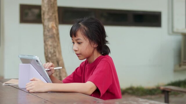 Asian kid using stylus pen on a digital tablet for e-learning at outdoor school table. Selective focus shows the young student engaging with technology for modern education and digital study concepts.