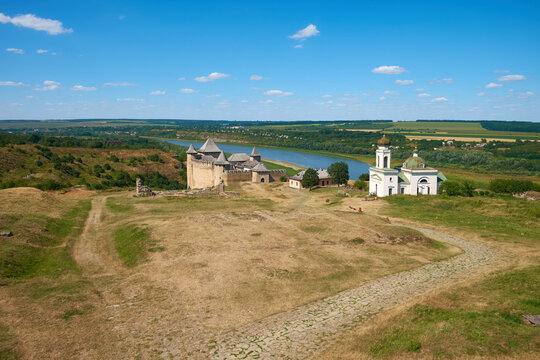Historic medieval fortress and church overlooking a winding river and rural landscape under a bright blue sky.