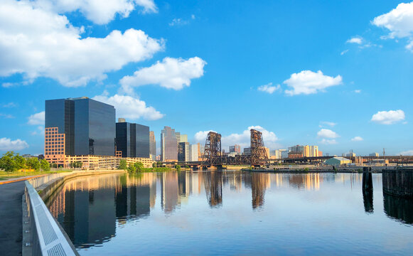 Panoramic view of Newark, New Jersey skyline reflecting on the Passaic River on a sunny day, featuring modern architecture and a historic vertical lift railway bridge under a blue sky.