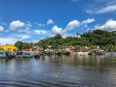 Scenic view of Santa Cruz Cabr&aacute;lia, Bahia, Brazil, featuring colorful boats, colonial houses along the river, lush hills, and a historic white church under a beautiful blue sky.