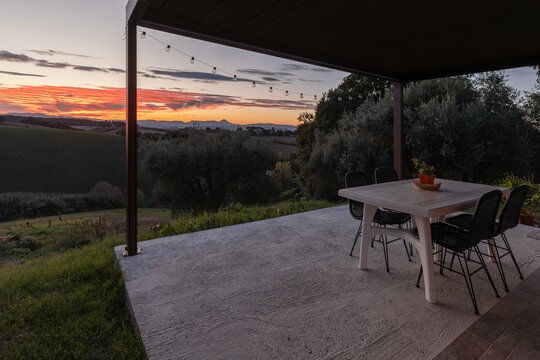 View of a patio with a table and chairs overlooking the rolling hills of the Marche region during a vibrant sunset with string lights in Petriolo, Marche, Italy.