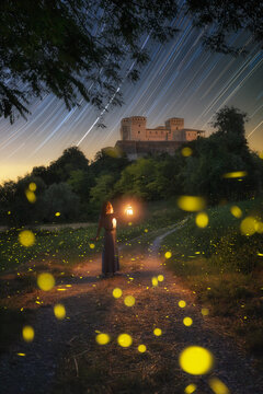 View of Torrechiara Castle at night with a woman holding a lantern on a path surrounded by glowing fireflies and star trails in the sky in Torrechiara, Emilia-Romagna, Italy.